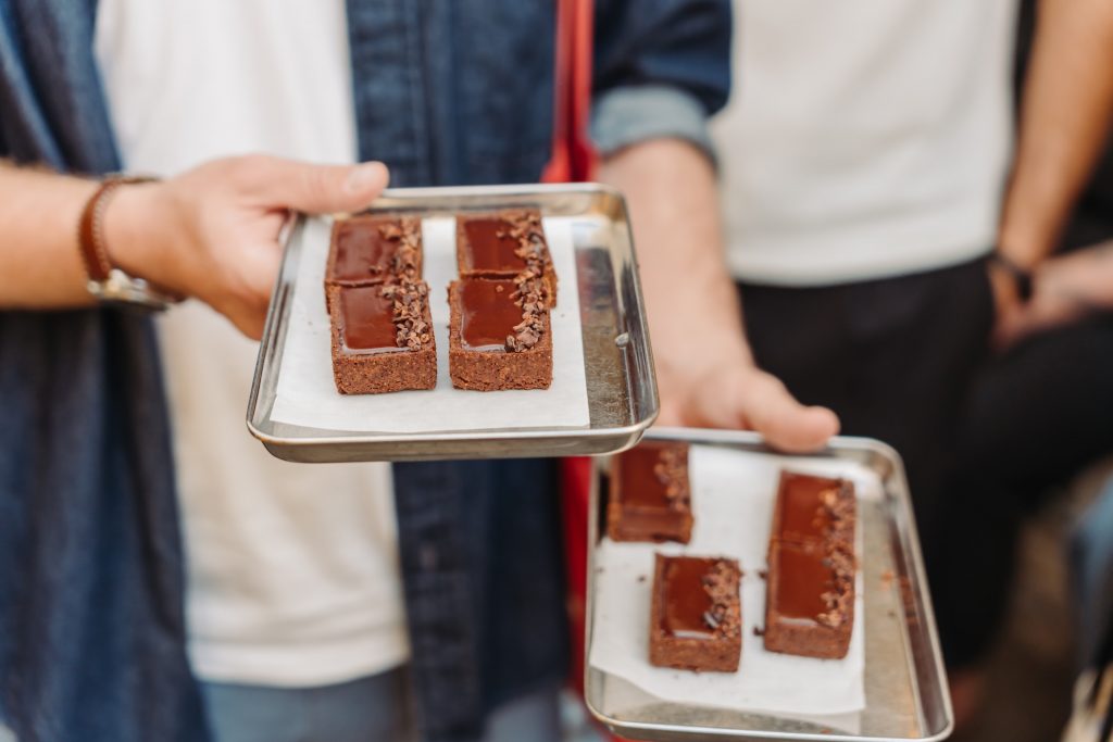 A person in a denim shirt offers two trays of brownies, each topped with chocolate or nuts, at a Paris bakery.