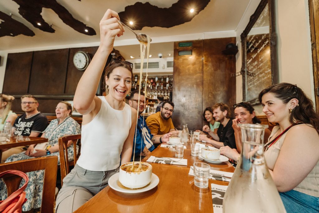 A smiling woman lifts a spoonful of cheesy French onion soup, cheese stretching, while chatting with friends at a busy Parisian restaurant.