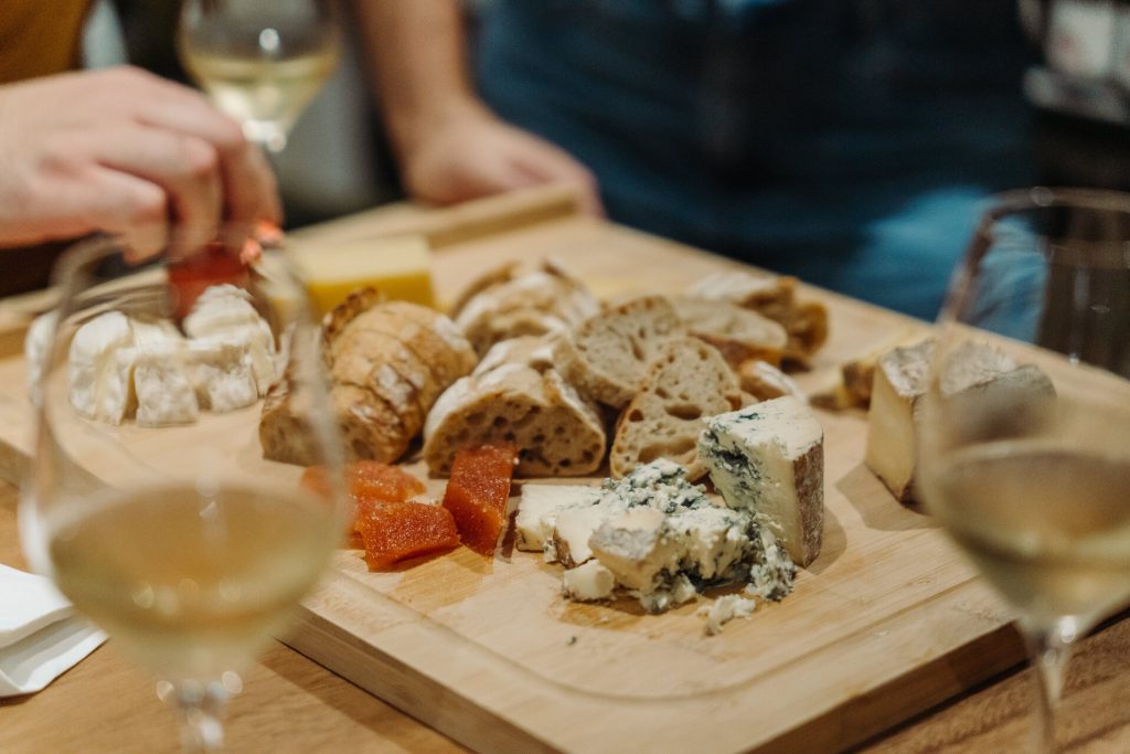 A hand reaches for cheese on a wooden board with assorted cheeses, sliced bread, fruit preserves, and three glasses of white wine.