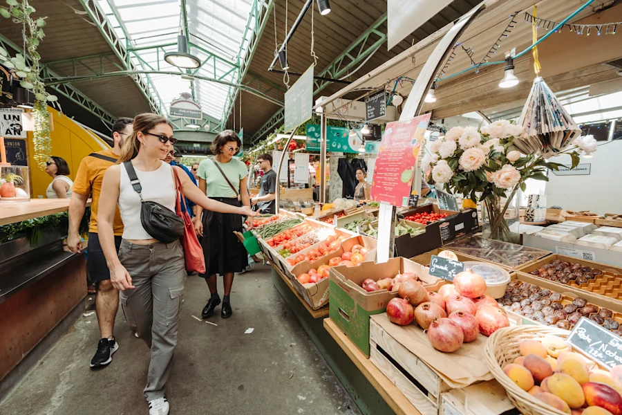 Shoppers browse colorful fruit and vegetable stalls under a bright, industrial ceiling at a Paris market; a woman points at tomatoes.