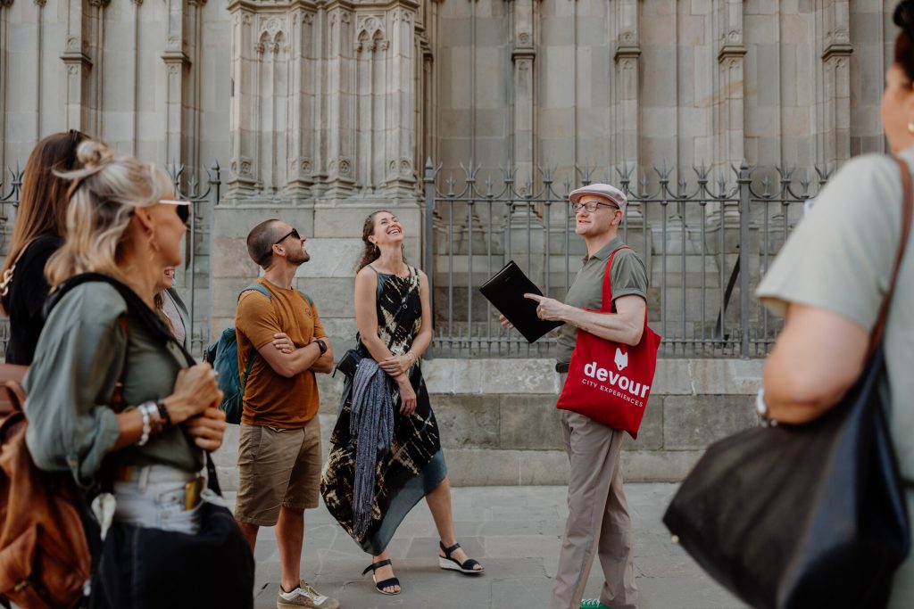A group listens to a tour guide with a red bag and notepad outside a historic stone building, likely in Barcelona.