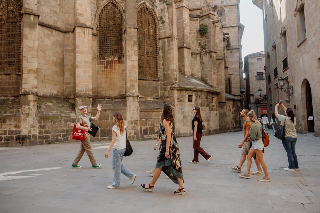 A tour guide leads attentive tourists with cameras past a historic stone building with arched windows on a sunny city street.