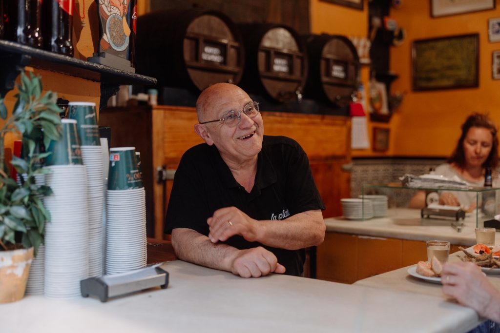 A smiling man in glasses chats behind a cozy cafe counter. Cups and bottles are stacked nearby. A woman eats in the background.