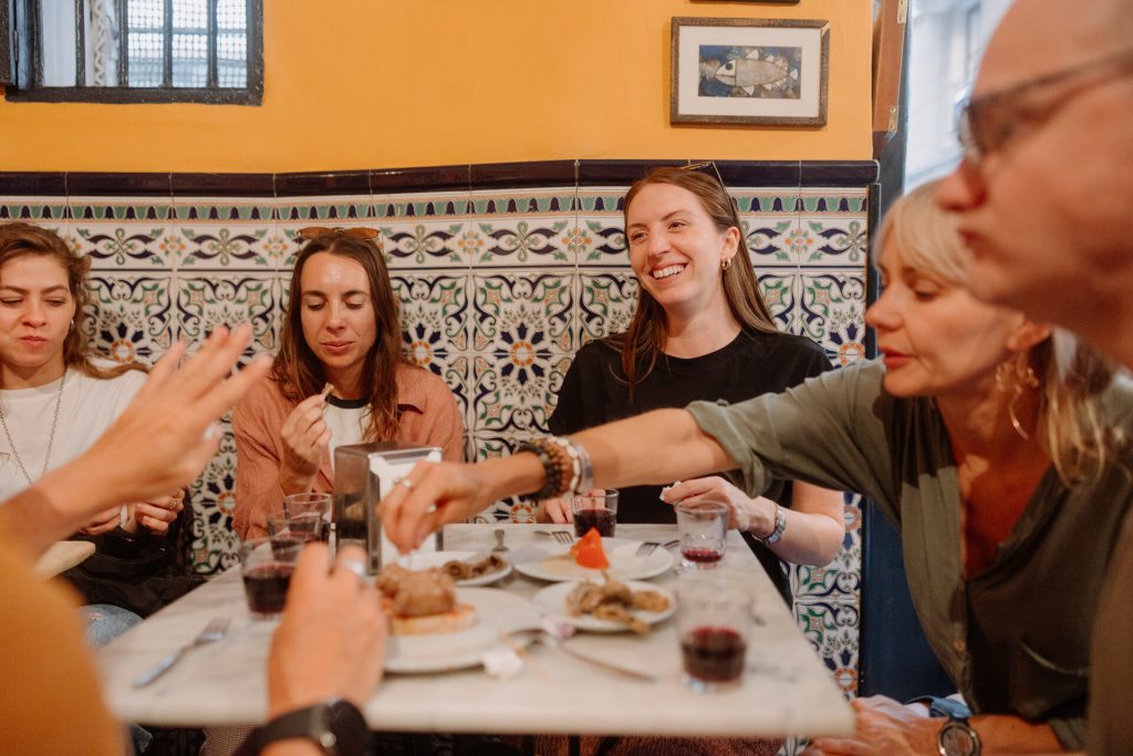 Several people share a meal and laugh together at a lively restaurant with vibrant tiled walls and framed artwork behind them.