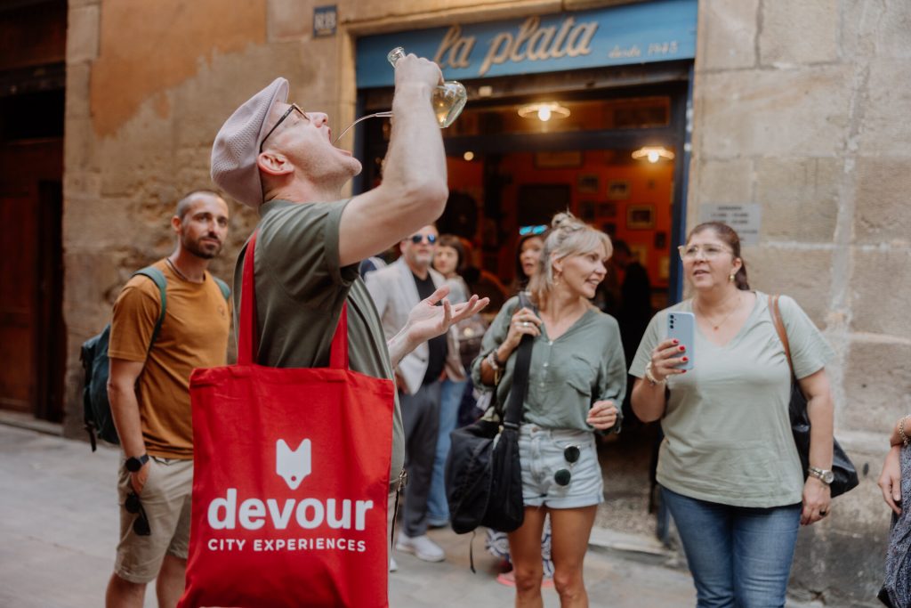 A man drinks wine from a glass pitcher on a rustic street in Spain, watched by several people near stone buildings.