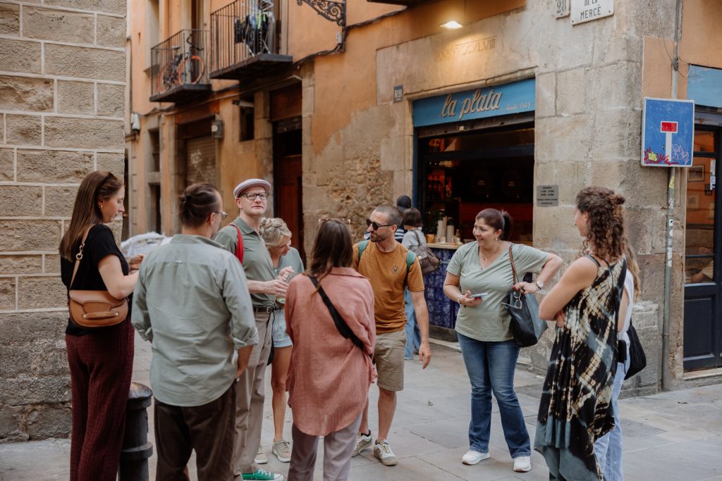 People chat near La Plata café in Barcelona; casually dressed, some listen to a woman with a notepad on the narrow street.