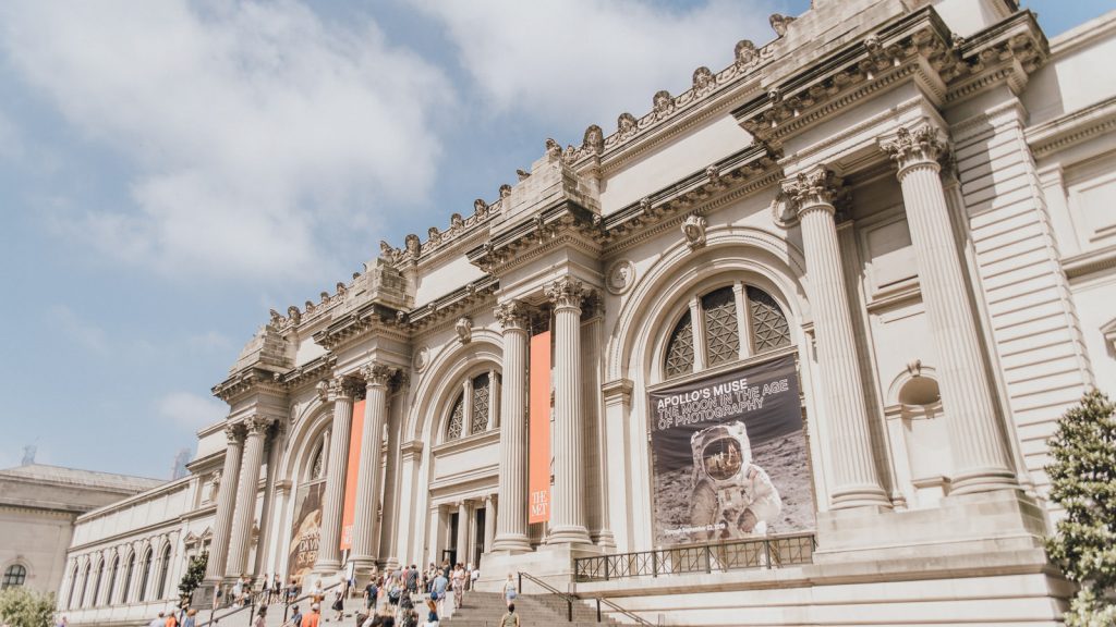 Visitors walk up the steps to the Metropolitan Museum of Art in New York City, passing grand columns and colorful banners.