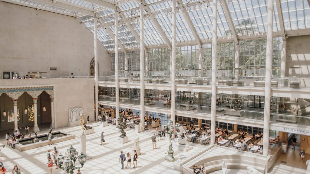 Visitors walk, sit, and explore exhibits in a bright museum atrium with a glass ceiling, sculptures, and a central fountain.