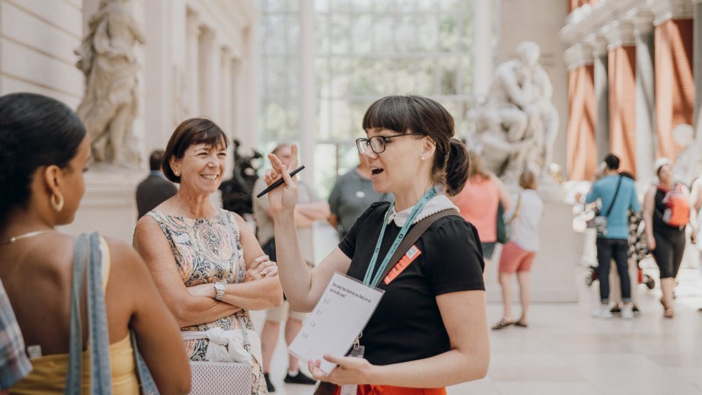 A woman with glasses and a lanyard leads a small group discussion in a bright museum gallery surrounded by statues.