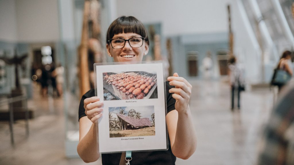 A smiling person with glasses displays a laminated sheet showing Betel Leaves and Rewa Canoe/Canoe House, inside a museum.