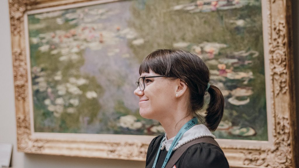 A woman with glasses and a ponytail looks at Monet’s “Water Lilies” at an art museum, standing quietly in profile.
