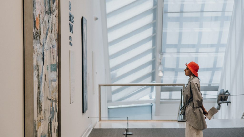 A person in a red hat and gray coat examines a large abstract painting by Mark Rothko in a well-lit contemporary gallery.