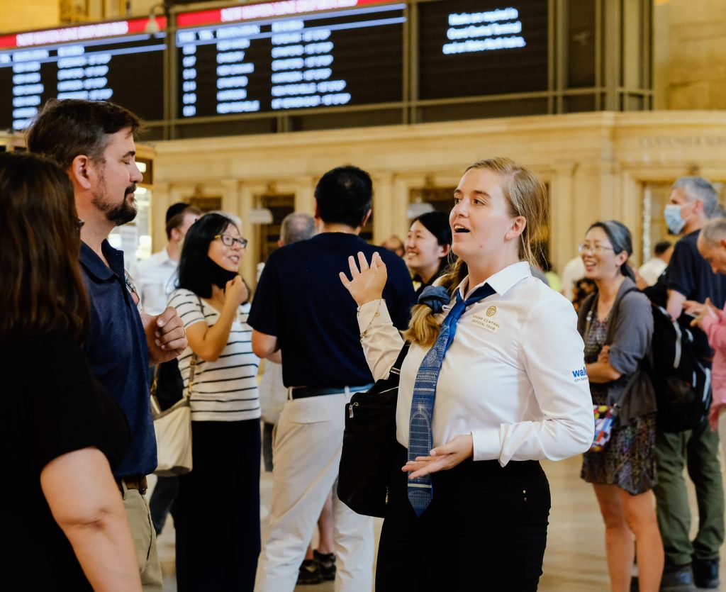 A woman in a white shirt and blue tie speaks to a man at a crowded train station near digital departure boards.