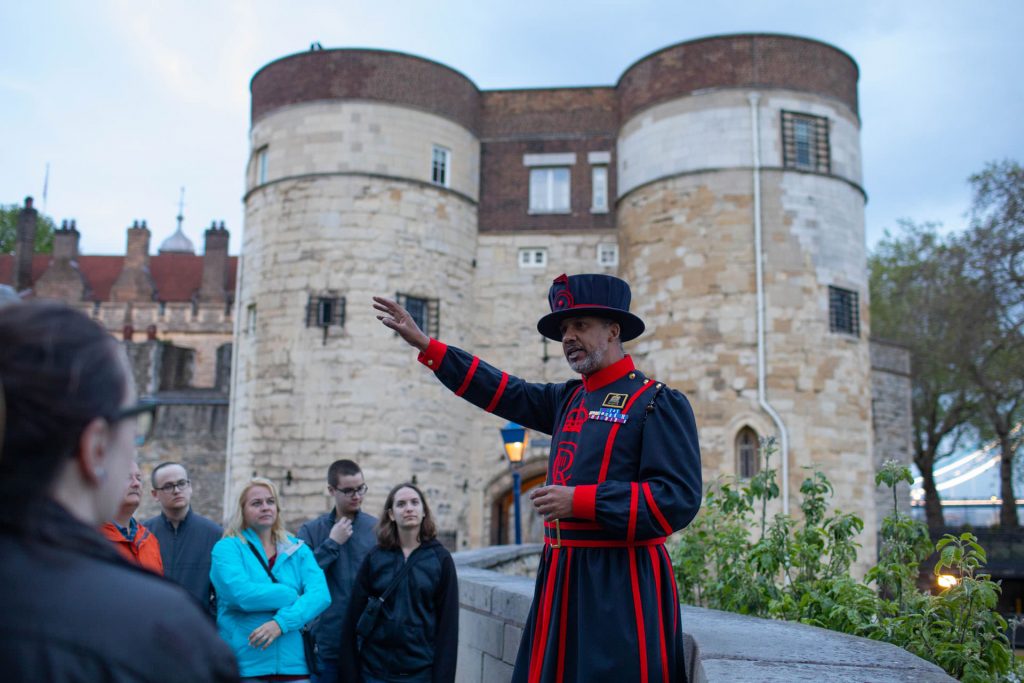 A Beefeater in traditional uniform leads attentive tourists outside the Tower of London as evening light casts shadows on ancient stone walls.