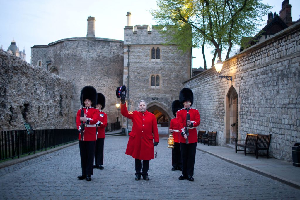 Four guards in red uniforms and bearskin hats stand in formation near a man raising his hat, outside London’s historic Tower.