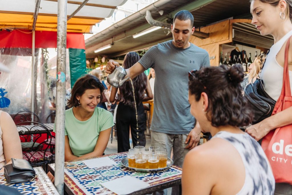 A man pours tea from a metal teapot into glasses as four women gather around a tiled table in a Parisian courtyard.