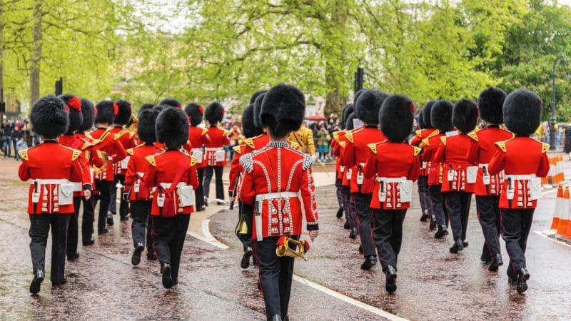 British guards in red uniforms and tall black hats march in formation on a wet road, watched by crowds and framed by trees.