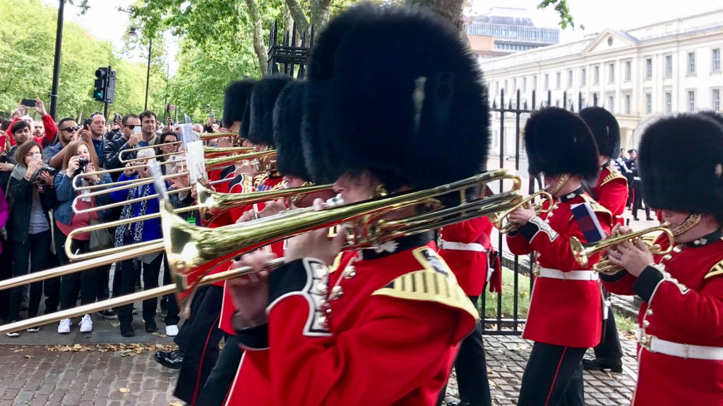 British guards in red uniforms and bearskin hats play trombones in a parade. Spectators watch behind a fence. Trees stand behind.
