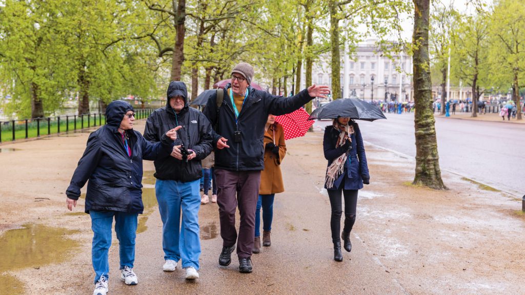 People walk on a tree-lined path in the rain, wearing jackets or carrying umbrellas. Wet puddles reflect the overcast sky.