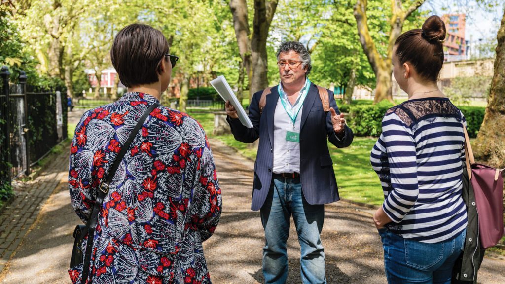 A man guides a walking tour in a sunny park, speaking with two attentive women among lush trees and green plants.