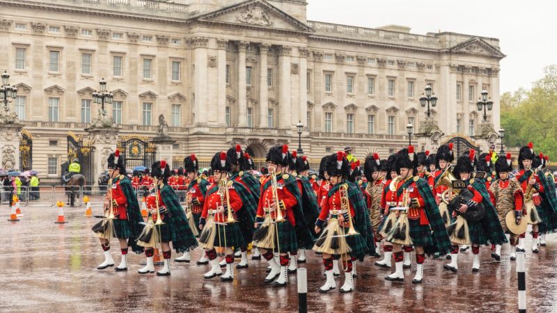 A Scottish marching band wearing kilts and feathered hats plays music outside Buckingham Palace during rainy weather.