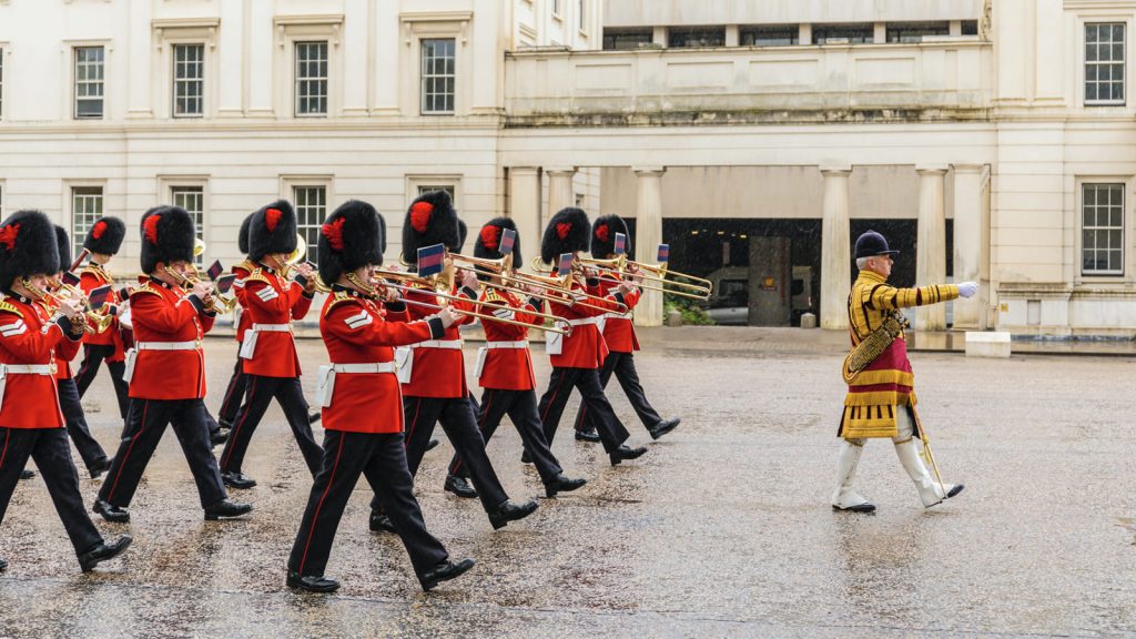 Soldiers in red uniforms and bearskin hats march in formation, playing brass instruments outside Buckingham Palace, London.