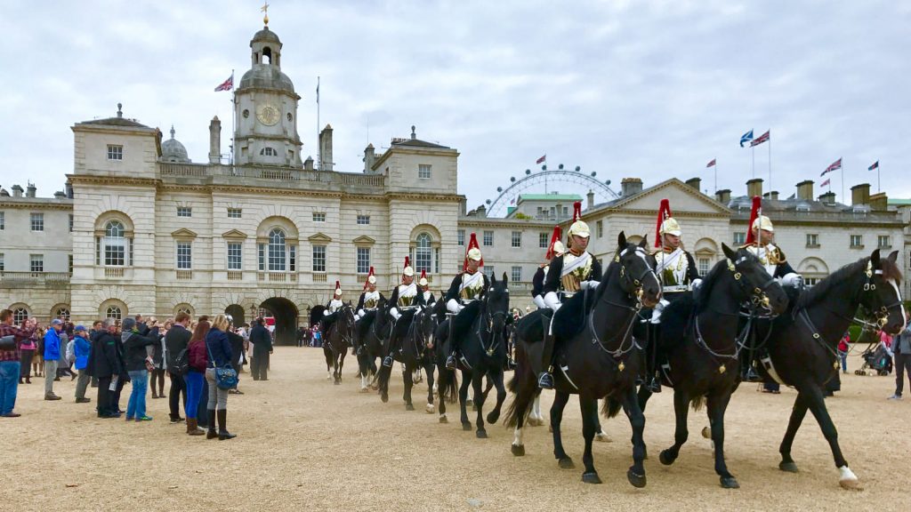 Mounted guards in ceremonial uniforms ride black horses past crowds near Horse Guards Parade, with the London Eye visible behind.