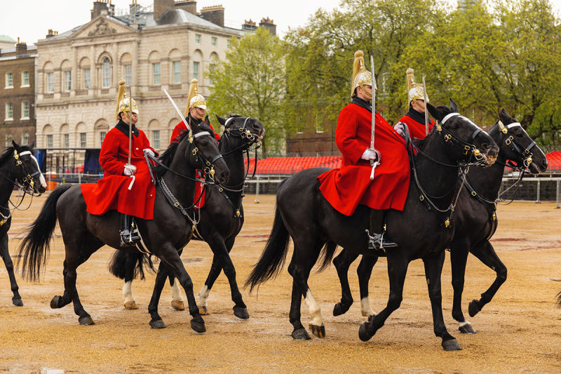Four soldiers in red uniforms and gold helmets ride black horses in formation. They pass a historic building, likely Horse Guards Parade, London.