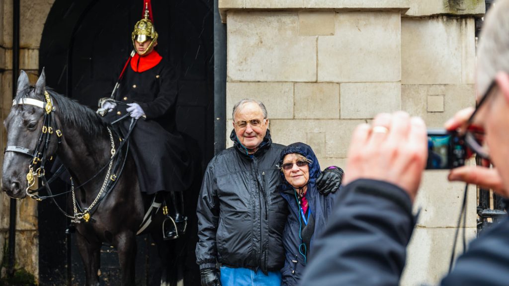 An older couple stands smiling in front of a Horse Guard on duty outside a stone building, while someone takes their picture.