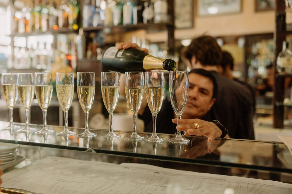 A bartender pours sparkling wine into several champagne glasses at a modern bar, with liquor bottles and mirrors behind him.