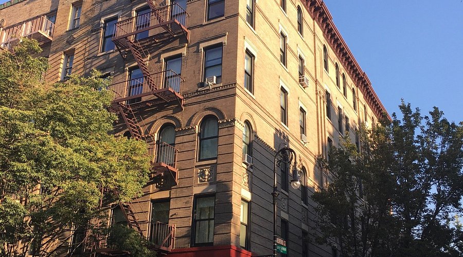 A tan brick apartment building with red fire escapes stands in Greenwich Village, partly shaded by leafy trees on a sunny day.