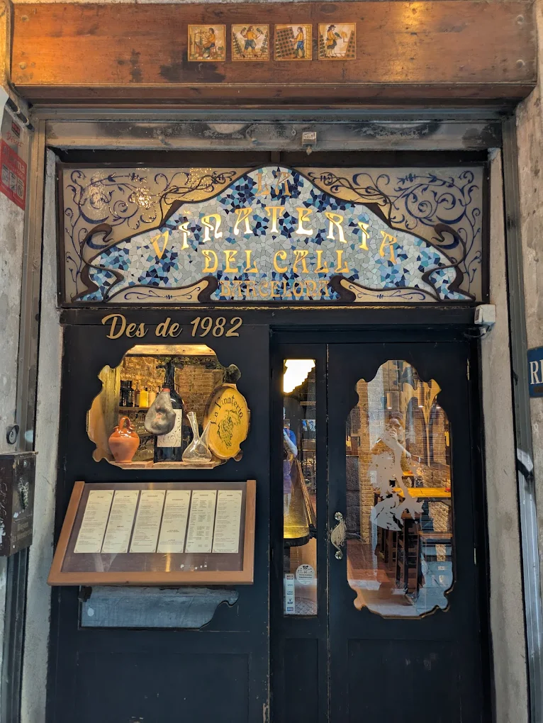 The entrance of Vini & Trine del Cali in Barcelona features ornate glass, gold-detailed doors, and menus by a sign from 1982.
