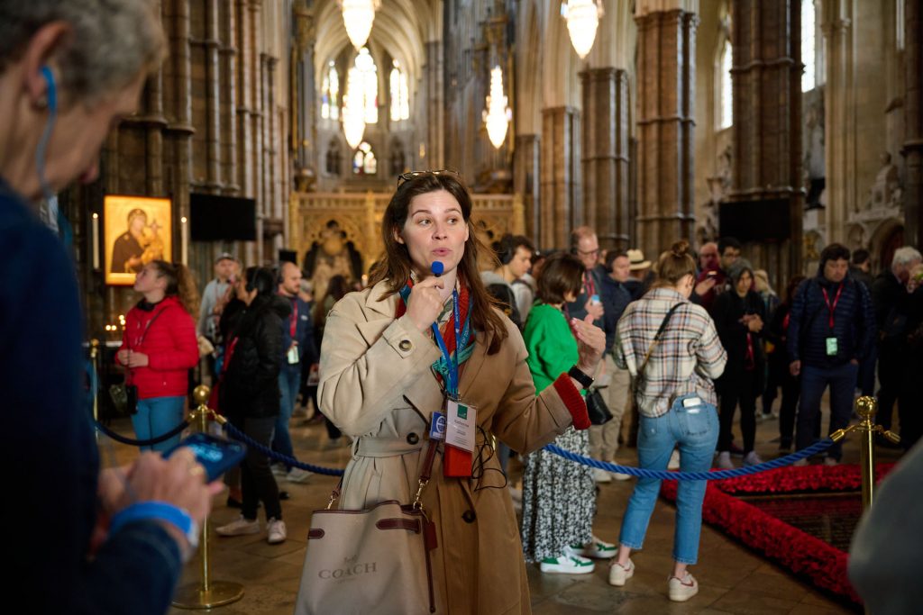 A woman guide in a beige coat leads a tour inside an ornate cathedral as visitors listen and explore the grand interior.