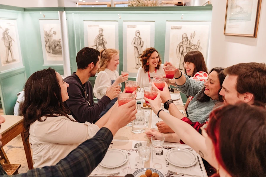 Four friends sit around a restaurant table with mint-green walls, smiling and toasting drinks beneath framed artwork.