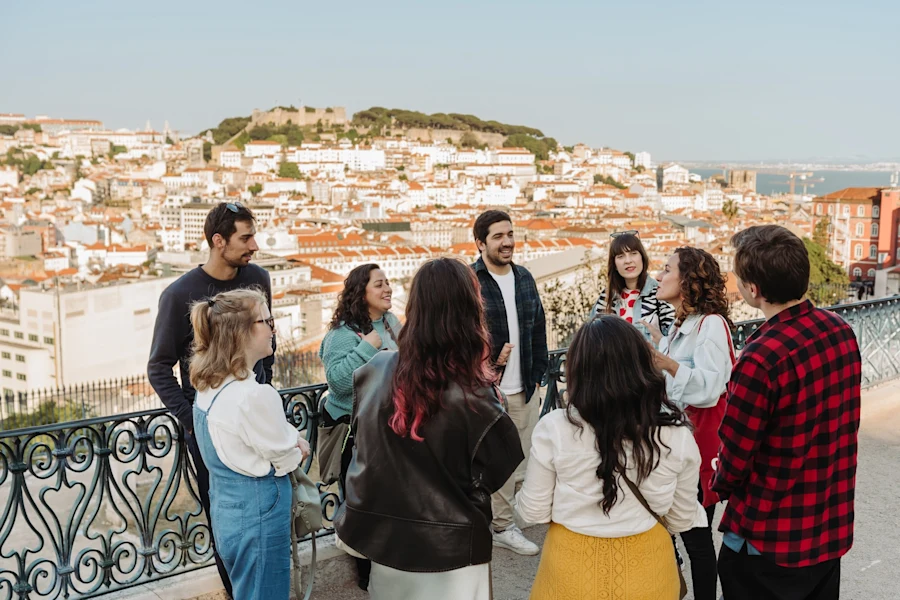 Several people stand in a circle, chatting and smiling outdoors. A sunlit cityscape and green hillside appear behind them.