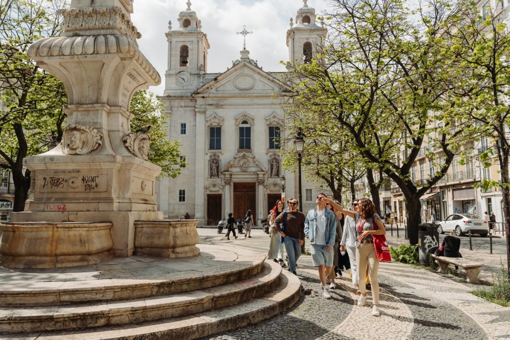 Several people stroll past a stone fountain in Lisbon’s sunny plaza, with leafy trees and a historic church behind them.