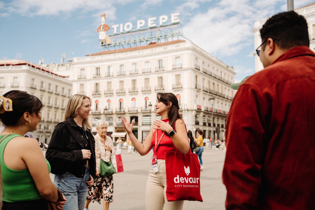 A tour guide with a red bag speaks to smiling tourists in Madrid’s Puerta del Sol, with the Tio Pepe sign behind them.