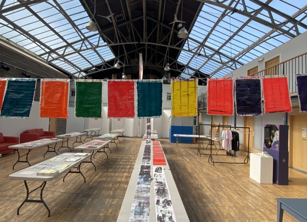 Colorful fabric panels hang above tables of books and art prints in a bright, glass-ceilinged Edinburgh gallery.