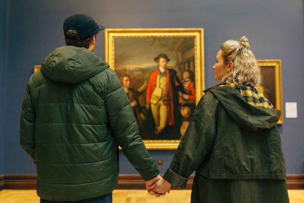 A couple in green jackets, holding hands, stand in Edinburgh museum facing a large framed painting by an unknown artist.
