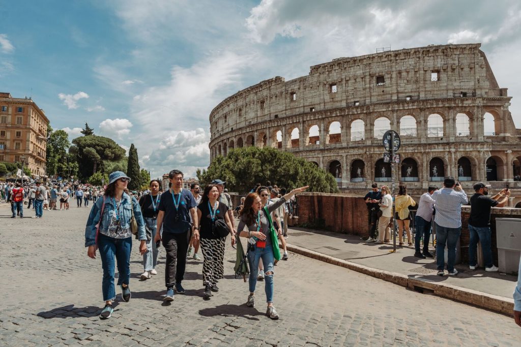 Tourists walk and talk with a tour guide in front of the Colosseum in Rome on a sunny day, with other visitors and old buildings nearby.
