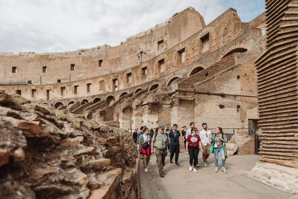 Tourists explore the ancient Colosseum in Rome, walking with a travel guide among arched stone walls and historic ruins under a partly cloudy sky.