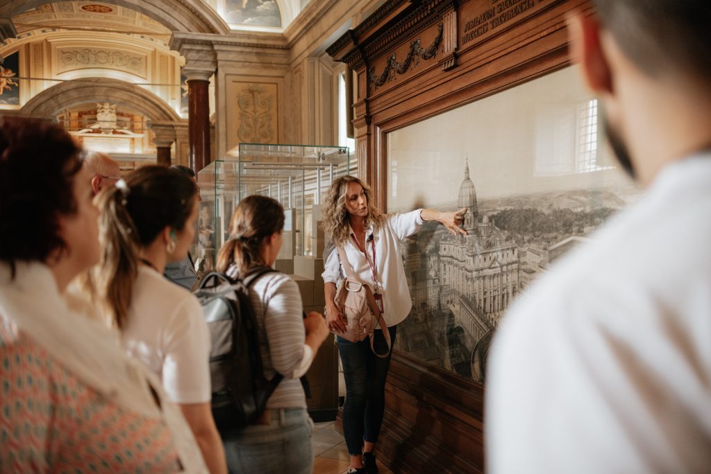 A tour guide gestures toward a large framed map as visitors listen in an ornate hall with arched ceilings and intricate decor.