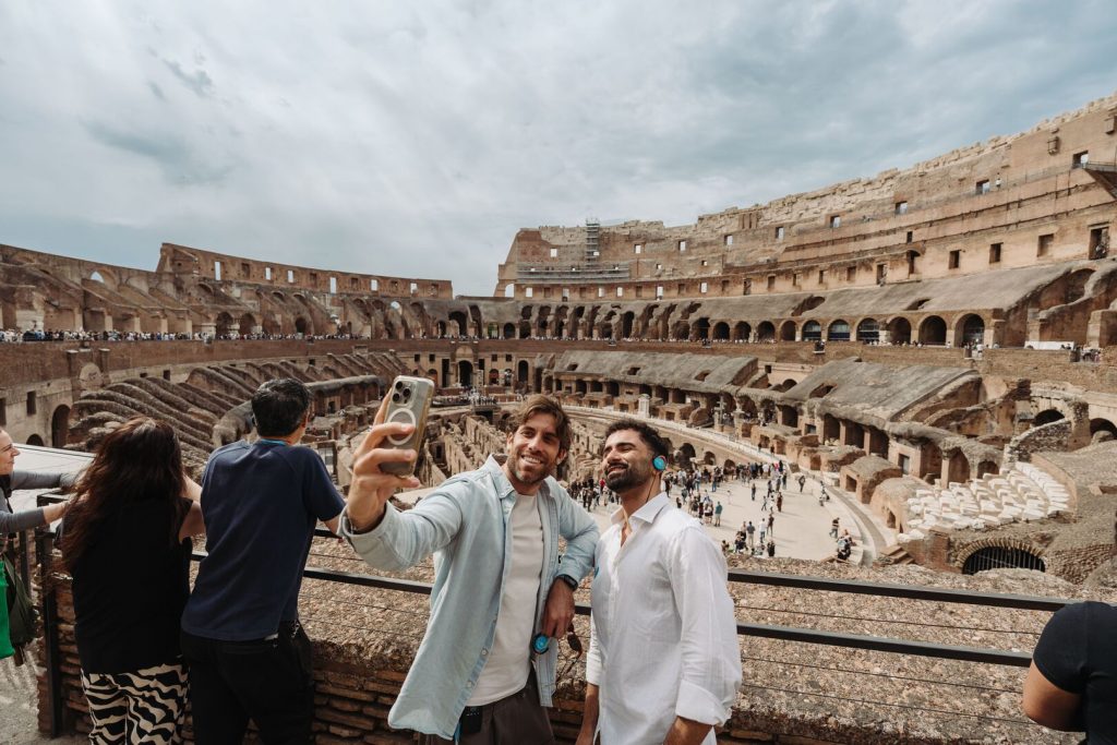 Two men smile while taking a selfie inside Rome’s Colosseum, surrounded by tourists and ancient stone walls beneath a cloudy sky.