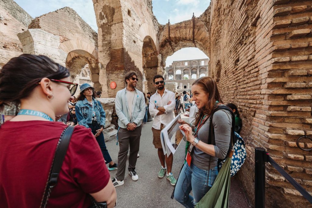 A tour guide in the Colosseum, Rome, shares photos with a group, who listen as ancient arches and brick walls rise around them.