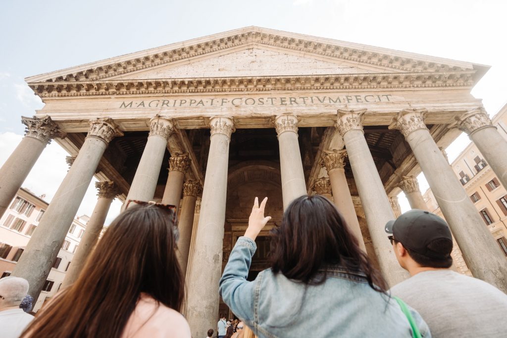 A travel guide and two tourists gaze up at the Pantheon’s grand columns in Rome. One flashes a peace sign. The sky is clear and blue.
