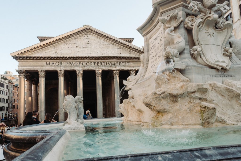 Water streams from the Fontana del Pantheon fountain as people gather beneath the columns of Rome’s ancient Pantheon temple.