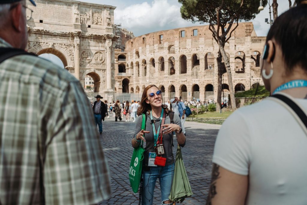 A cheerful tour guide leads a group in Rome, gesturing in front of the Colosseum and Arch of Constantine amid ancient ruins.