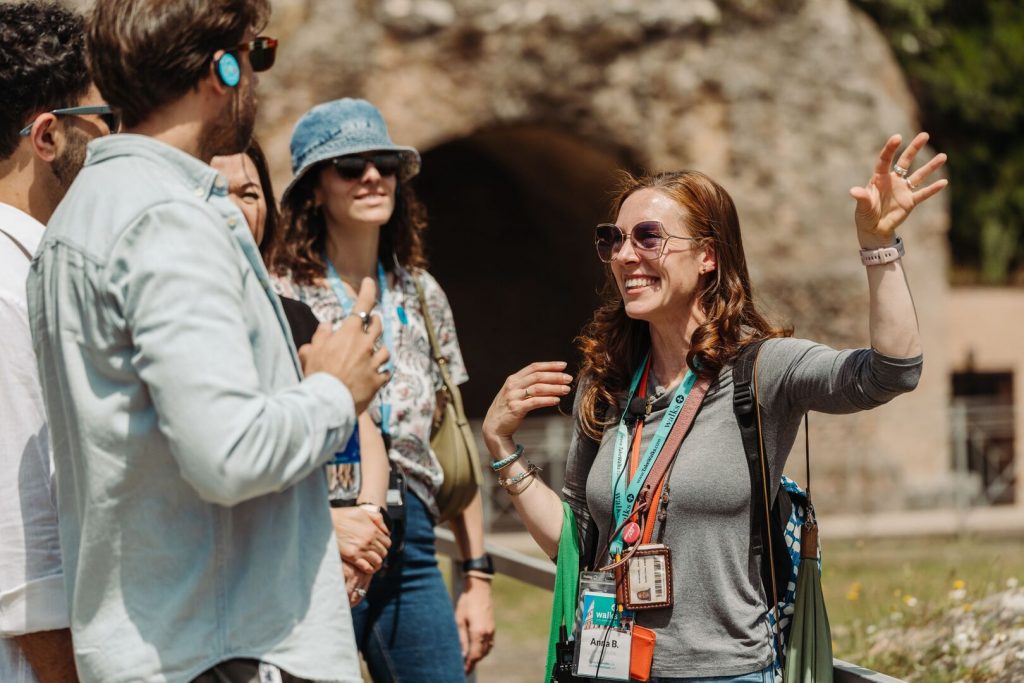 A smiling woman with sunglasses enthusiastically leads a small tour group beside an ancient stone structure outdoors, possibly historic ruins.