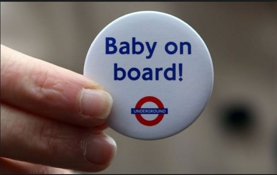 A person holds a white "Baby on board!" badge featuring the London Underground logo, likely for use while traveling on the Tube.