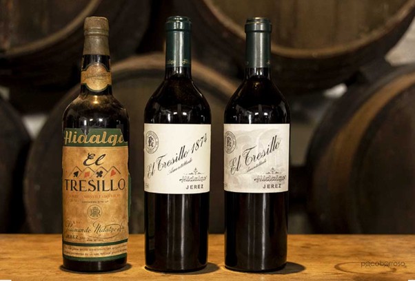Three El Tresillo sherry bottles are placed on a wooden table in Jerez, with blurred aging barrels visible behind them.